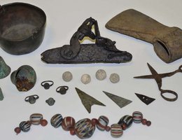 European goods manufactured for trade with Native Americans, including glass beads, metal rings, lead musket balls, part of a flintlock musket, metal scissors, a metal hatchet, a metal pot, and arrow points fashioned from metal objects